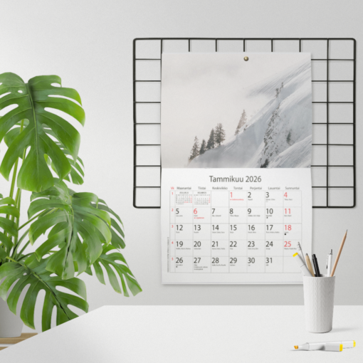 Seinäkalenteri vuodelle 2026 esillä modernissa työtilassa. Kuukauden kuvana oma valokuva: talvinen vuoristomaisema sumun keskellä.