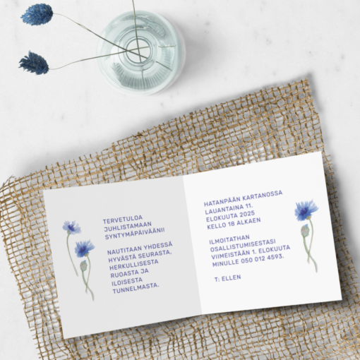 Kuvassa kaksiosainen syntymäpäiväkutsukortti, jossa on sisäsivuille painettuna teksti ja sinisiä ruiskaunokkikuvituksia. Kortti lepää vaalealla pöydällä, jonka päällä on koristeena verkko sekä lasinen maljakko, jossa on kuivatut kukat.