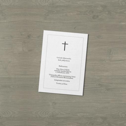 Hautajaiskutsu, jossa risti ja suruviesti, aseteltu harmaalle puupinnalle vinosti.
