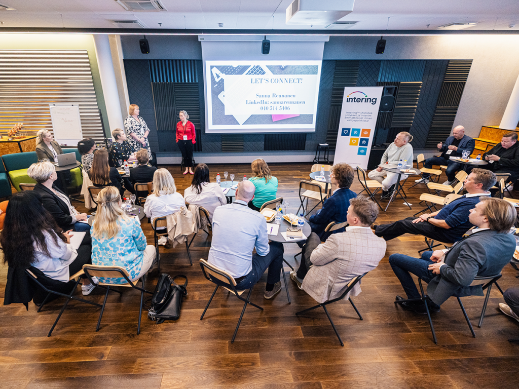 Intering-tapahtuman osallistujia kuuntelemassa esiintyjiä salissa, taustalla suuri valkokangas.