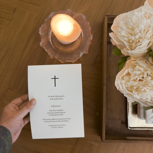 Hautajaiskutsu kädessä pöydän ääressä kynttilän ja kukkien vieressä.