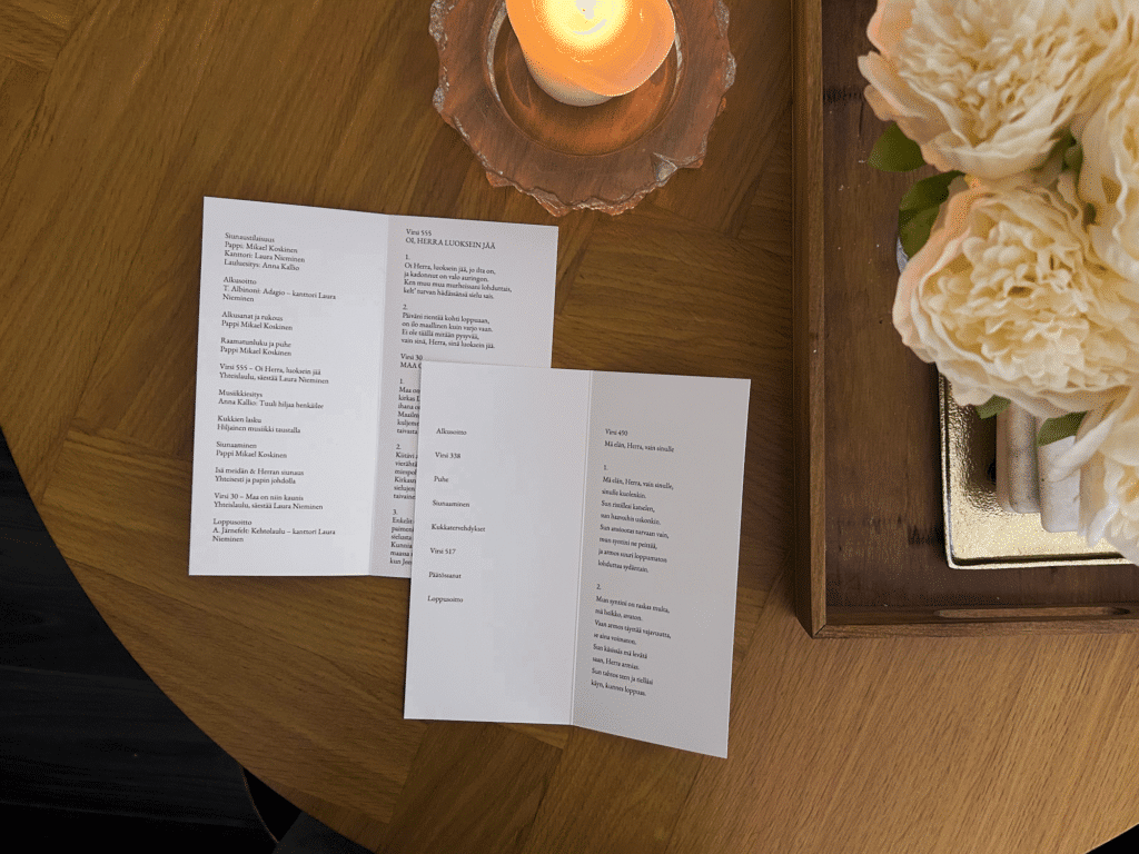 Pitkämallinen hautajaisohjelma avattuna pöydällä kynttilän vieressä.