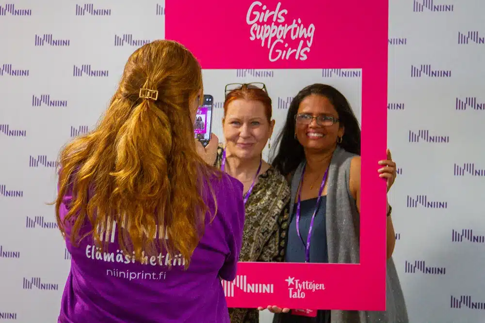 Kaksi osallistujaa poseeraa pinkin Girls Supporting Girls -kehyksen sisällä samalla kun heitä kuvataan.